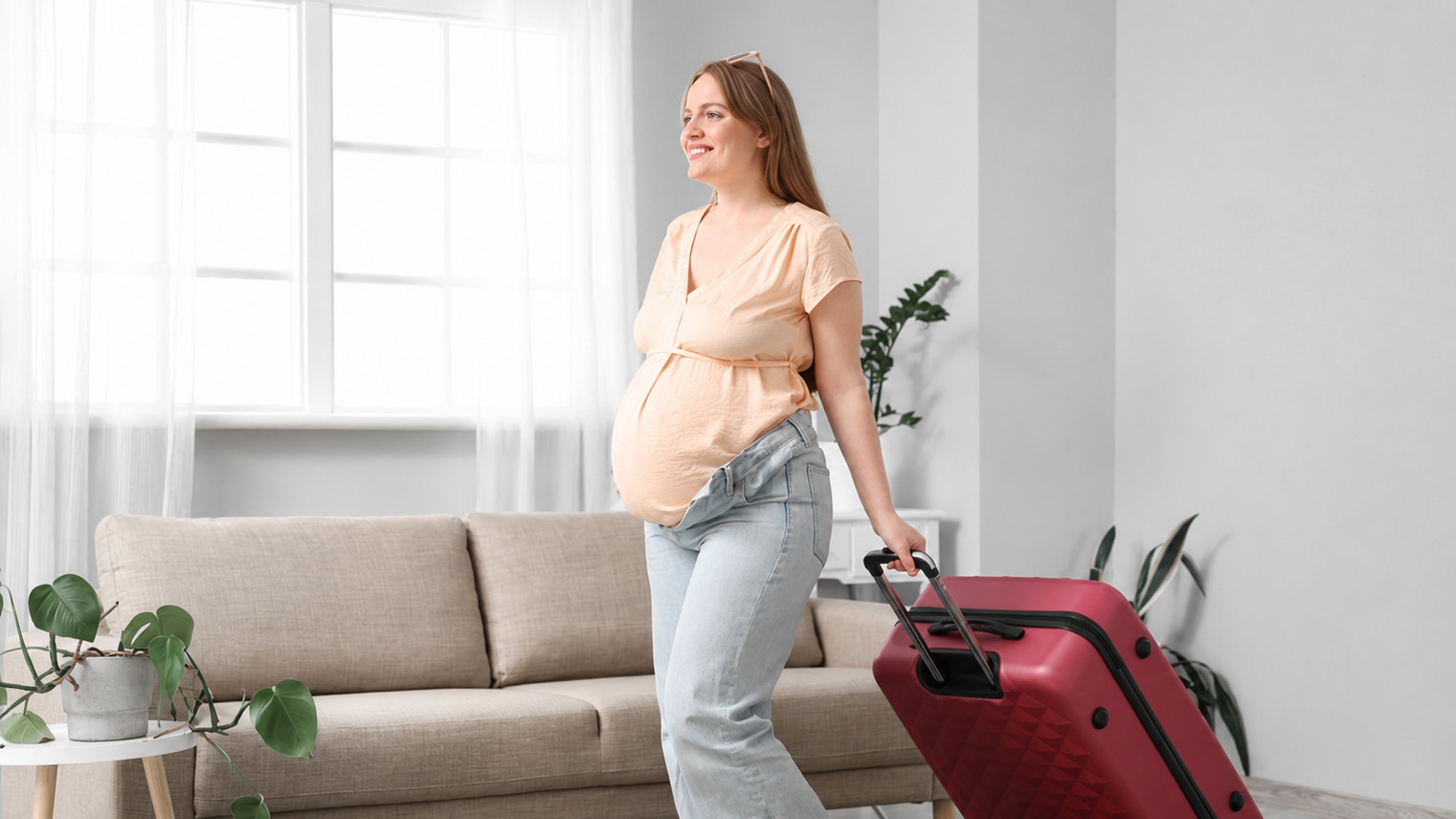 Pregnant woman standing with a travel suitcase at home, illustrating overseas childbirth as part of citizenship planning